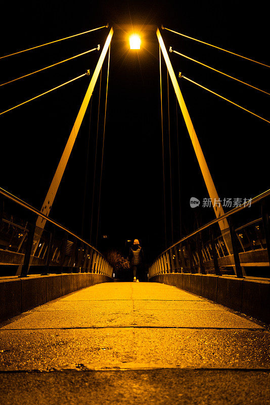 夜景中对称的斜拉桥用钢索形成引导线，一个女孩背着她走着。一盏街灯发出淡黄的光，照亮了整个场景。