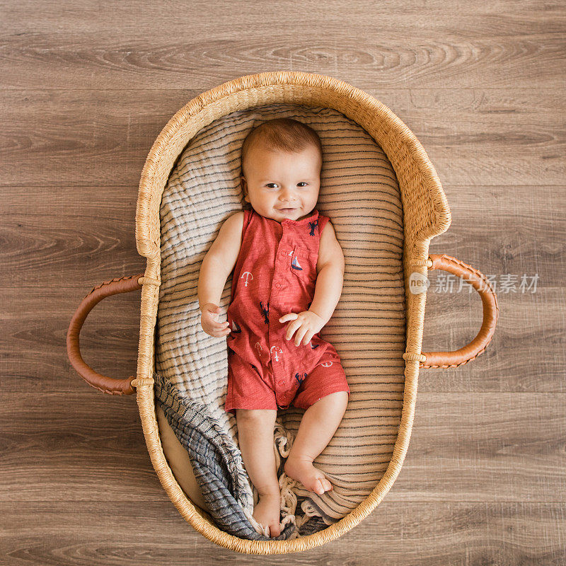 一个18周大的微笑小男孩穿着红色航海服躺在一个舒适的奶油色条纹棉毯在海草摩西篮子