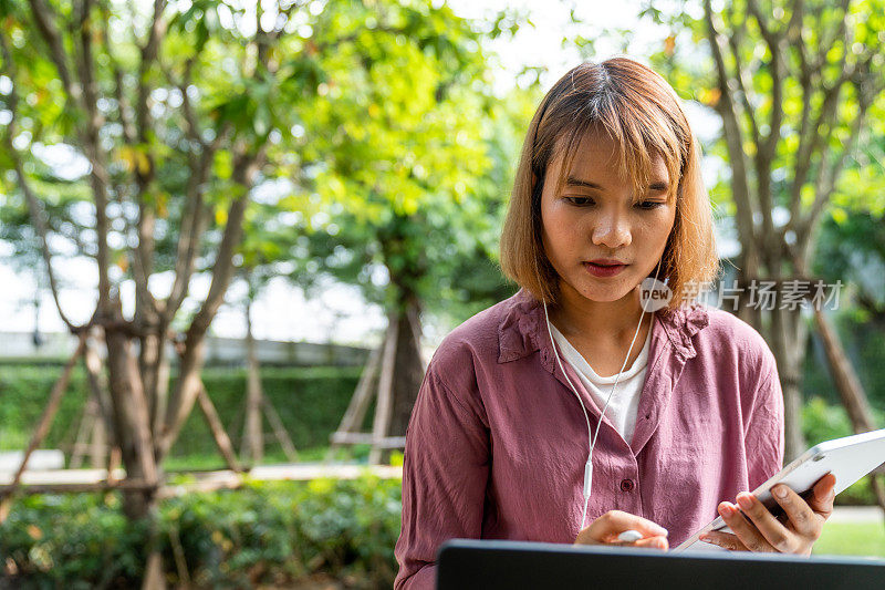 小女孩戴着耳机，用笔记本电脑上网学习，坐在公园里在平板电脑上写字。