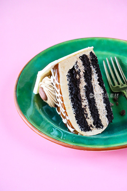 咖啡和大理石巧克力蛋糕片的图像，三层蛋糕被咖啡奶油在翠绿色的盘子上用叉子分开，管道奶油玫瑰，巧克力咖啡豆，豪华巧克力蛋糕，巧克力刨花，粉色背景