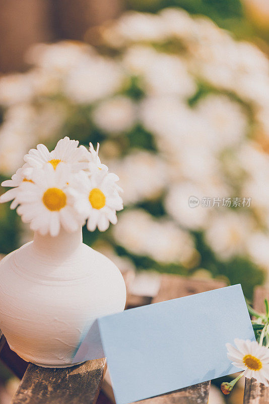 一个白色的小花瓶，上面插着白色的雏菊，一张蓝色的空纸，作为你复活节、春夏短信的原型，放在一张乡村小桌子上