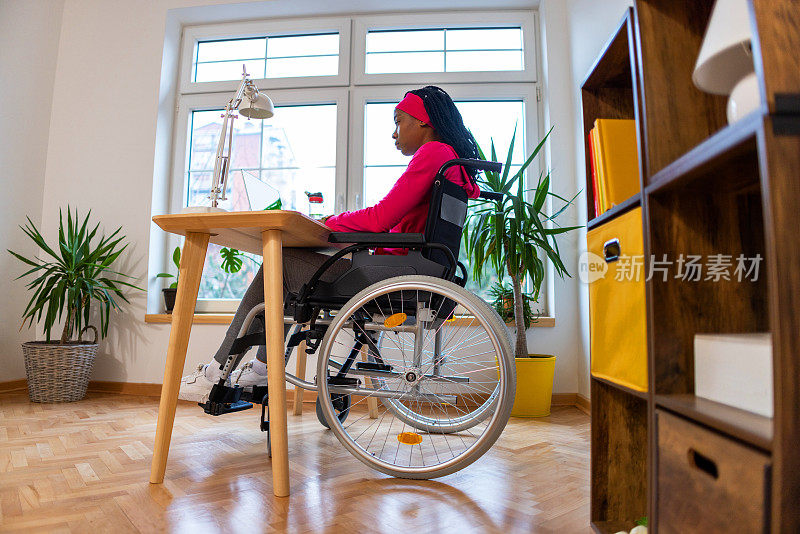 黑人妇女坐在轮椅上用笔记本电脑工作，在她的家庭办公室