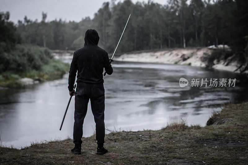 在雨中，一个黑衣男子背对着镜头站在河边的森林里，手里拿着武士刀。黄昏时分，一位英俊的男子手持武士刀。