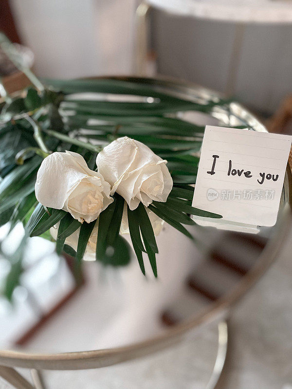 这是一幅静物画，玻璃桌上放着一朵白玫瑰，一张纸上手写着“我爱你”