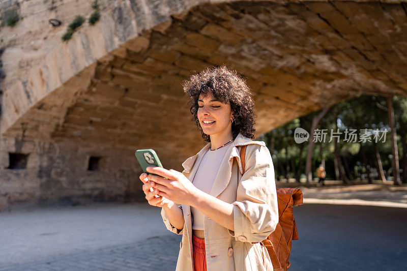 微笑的无忧无虑的年轻女子站在户外，用手机技术探索外国城市，在智能手机在线地图上寻找位置。