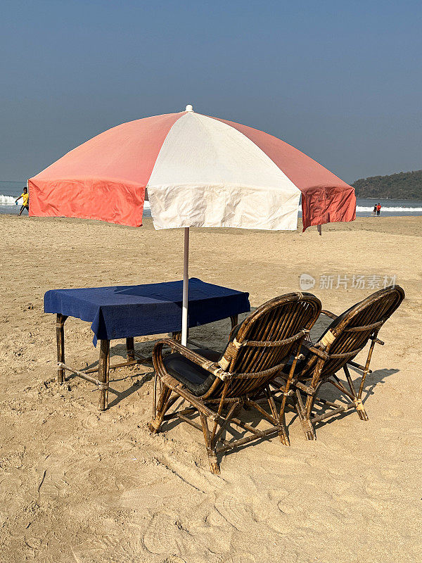 金色沙滩上蓝色桌布覆盖的餐桌，竹椅和条纹遮阳伞，沙滩上游客的露天用餐区，蓝天和大海的背景，重点在前景