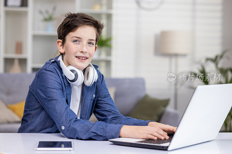 脖子上戴着耳机的微笑少年，坐在明亮的房间里的笔记本电脑前，敲击键盘，学习，看视频，在线聊天，看着相机