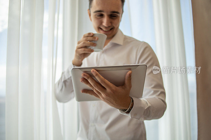 一个穿着白衬衫的男人站在酒店房间的窗边，手里拿着一杯咖啡和一台平板电脑