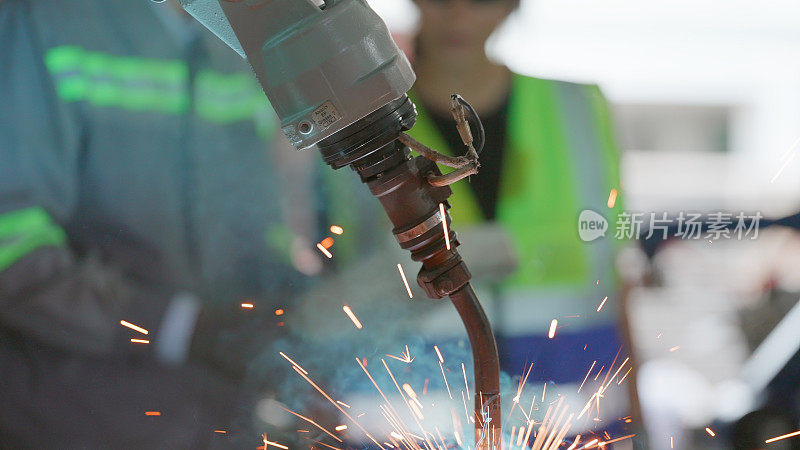 用于工厂生产技术的智能工业机械臂。展示了工业自动化制造过程，机器人手臂焊接工件在后台，一名工人使用控制面板控制机器人的操作。
