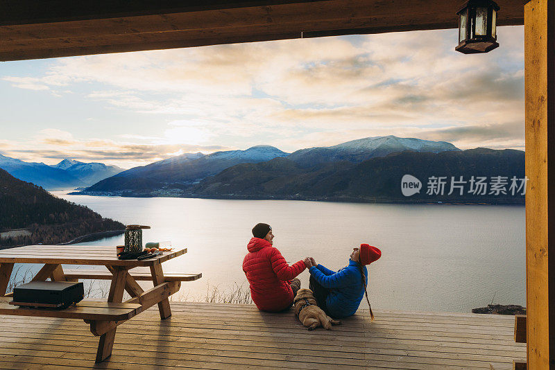 在挪威的日落时分，幸福的夫妇和狗在避难所欣赏冬季峡湾的美景