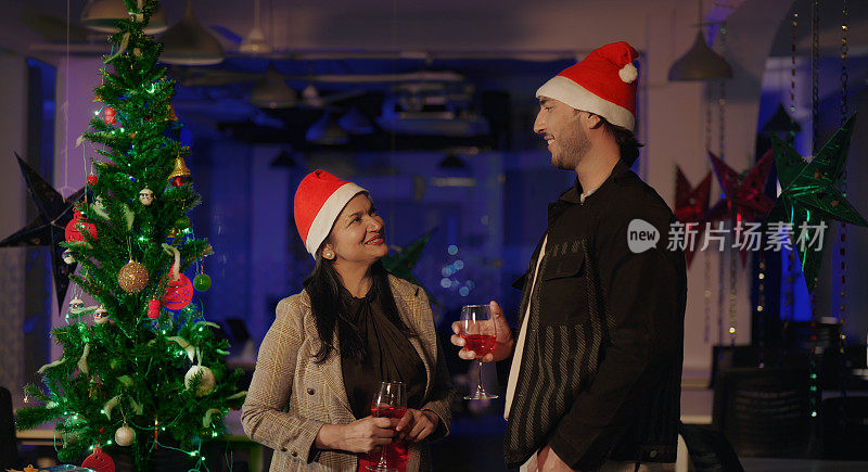 两位戴着圣诞帽的印度同事站在室内办公室，端着杯子，喝着果汁，一起享受夜晚的2024年新年派对。快乐微笑的漂亮女性年轻男性做有趣的八卦