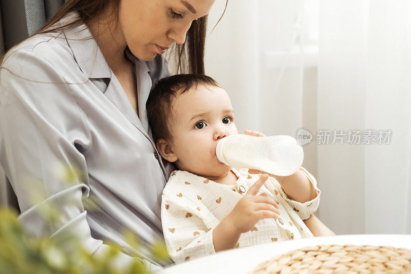 孩子和妈妈坐在厨房的椅子上，手里拿着一个瓶子，喝着里面的婴儿配方奶粉。