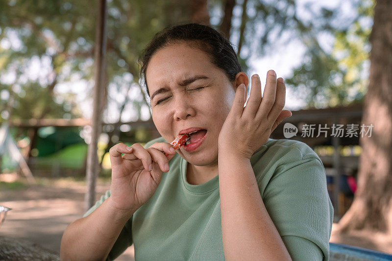 一名穿着休闲服装的成年亚洲女性坐在当地的一家餐馆里，吃着僵硬的食物，由于牙痛的症状而感到疼痛，需要去看牙医。