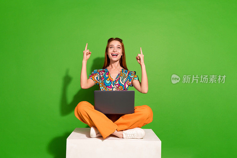 漂亮的年轻女孩的全身肖像坐在讲台上笔记本电脑直接手指向上空的空间穿顶隔离在明亮的绿色背景