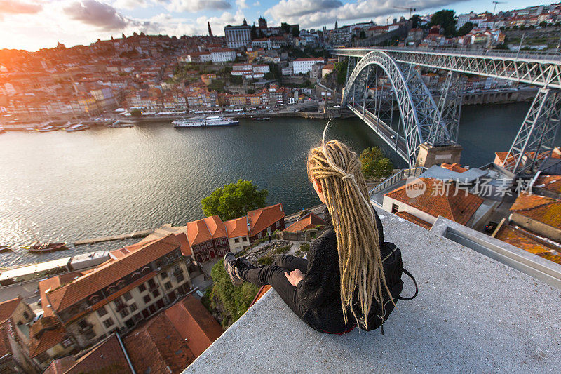 在葡萄牙波尔图，一个金发长发绺的年轻美丽的女人在杜罗河对岸的多姆·路易斯桥对面的观景平台上与夕阳相遇。
