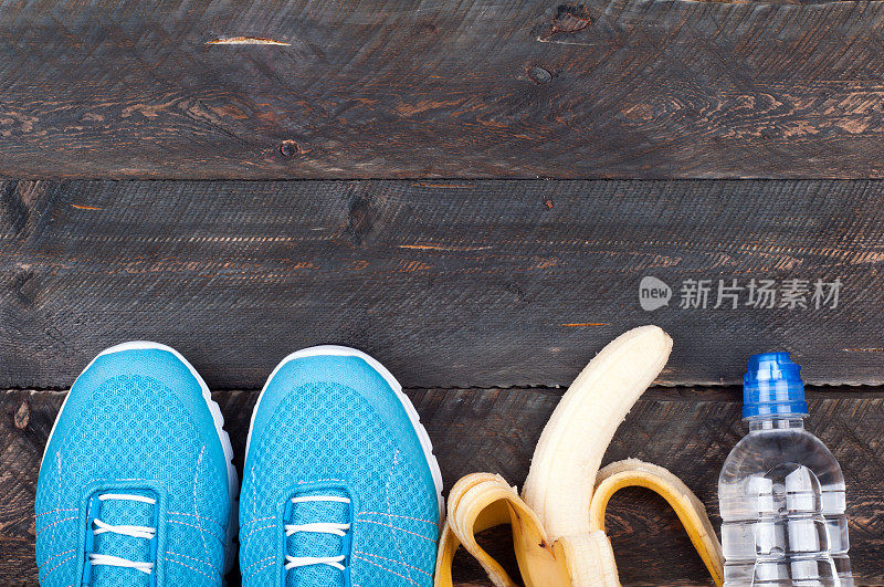 运动设备。运动鞋，香蕉和瓶装水在深色木制背景。积极健康的生活理念背景与复制空间。俯视图