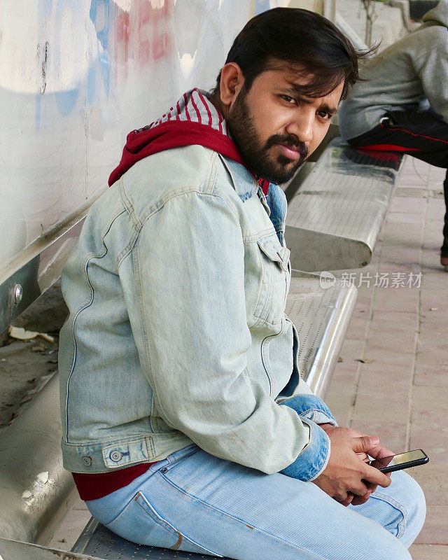 在印度新德里，一名年轻的印度男子留着胡子，拿着手机在户外给朋友发短信