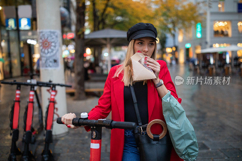 一名年轻女子在人行步行区边吃边扶着滑板车