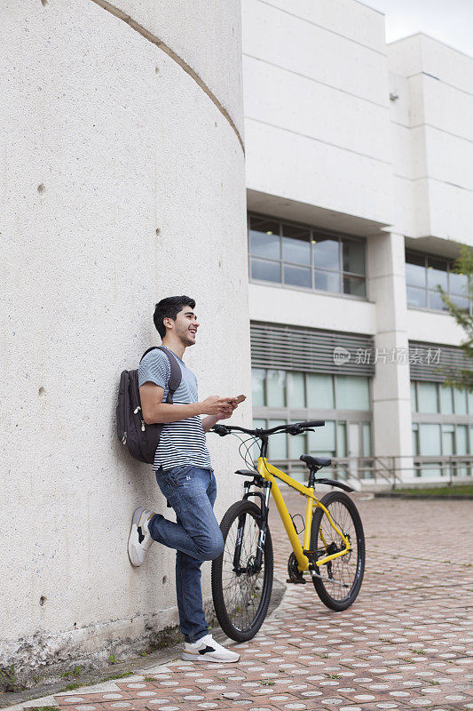 24岁的拉丁裔男子穿着很随意，身边停着一辆摩托车，在一个阳光明媚的日子里，他倚在学校外的一面墙上查看手机