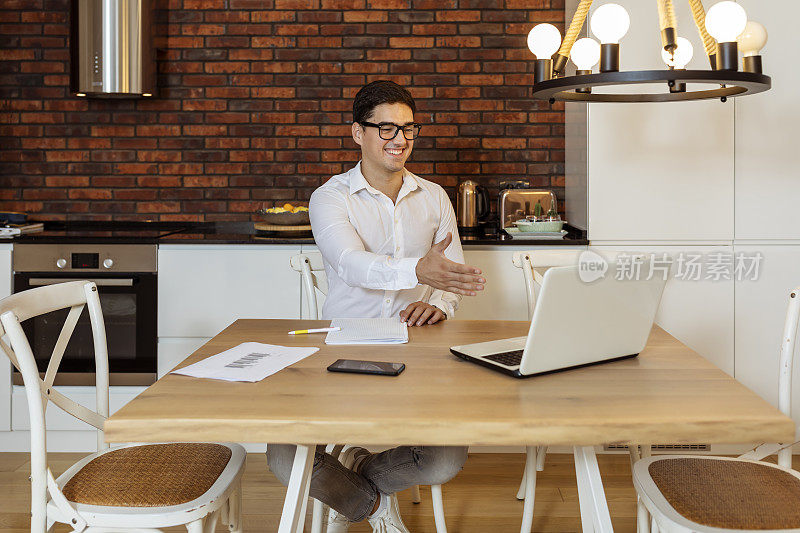 肖像快乐的年轻人戴着眼镜坐着，看着笔记本电脑屏幕上的视频通话和握手