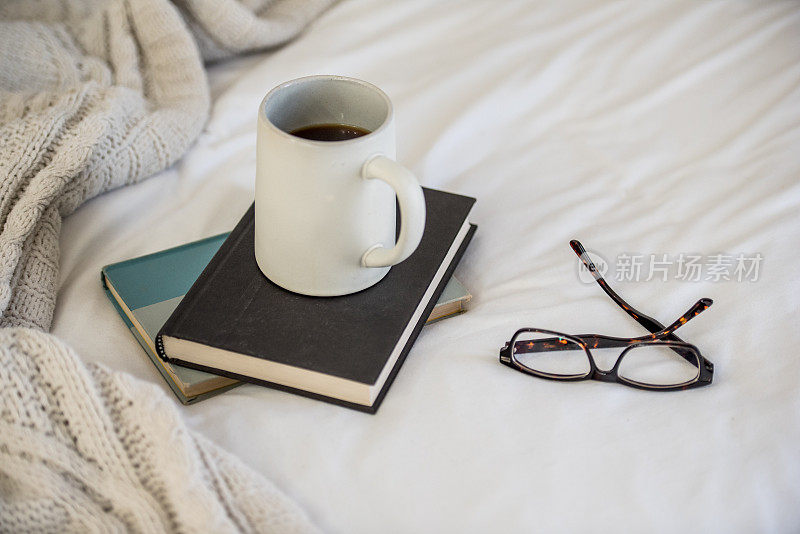 一张舒适的居家照片，一张床上的毯子上放着马克杯、书和眼镜。