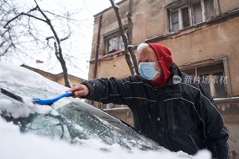 由于COVID-19疫情爆发，一位积极工作的老人戴着防护口罩，用塑料刮冰器和扫把清除汽车挡风玻璃上的积雪
