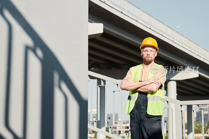 自信的男性建筑工人的肖像，戴着安全护目镜和安全帽，双臂交叉靠在桥上