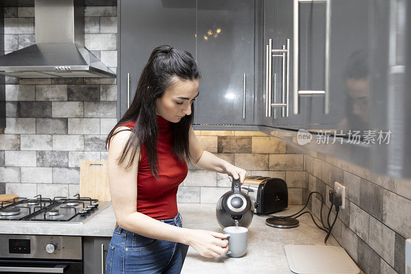 一名女大学生在厨房柜台边泡茶，一边把水壶里的水倒进杯子里