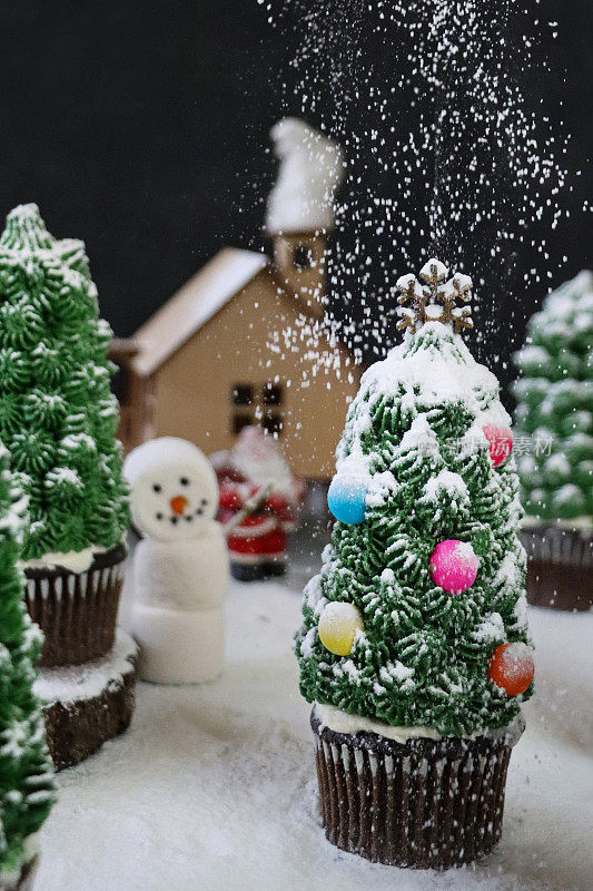 特写图片是批量自制的，圣诞树设计的巧克力纸杯蛋糕放在纸杯蛋糕盒里，展示在夜晚针叶林的场景中，有飘落的冰霜雪粉，有圣诞老人和棉花糖雪人的模型房子