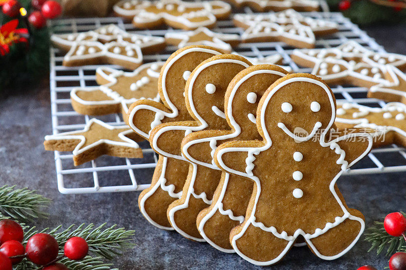 近距离拍摄的姜饼人饼干，除了饼干冷却架外，还装饰着白色的皇家糖霜，云杉针和红莓，发光的小圣诞树上的仙女灯，黑森包底，灰色背景，家庭烘焙