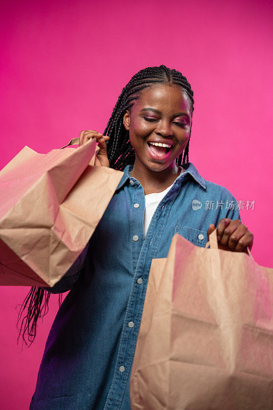 一个笑着的皮肤黝黑的年轻女孩，手里拿着大减价时买的购物袋，背景是亮粉色