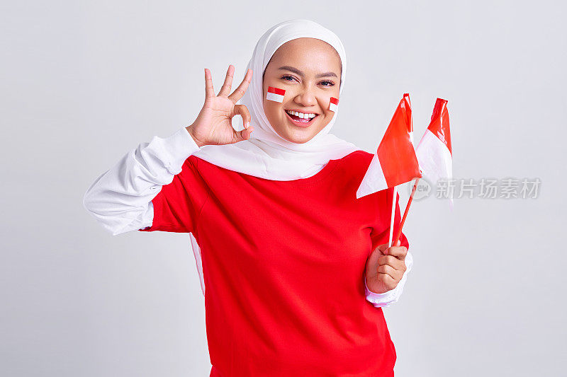 微笑的年轻亚洲穆斯林妇女穿着红白t恤，举着印度尼西亚国旗庆祝8月17日印度尼西亚独立日，并在白色背景上显示okay手势孤立