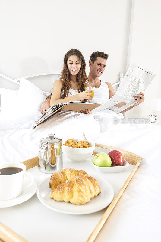 一对夫妇躺在床上，手里拿着报纸和早餐盘