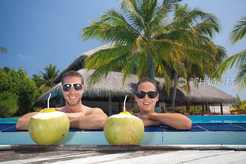 年轻夫妇与新鲜的绿色饮用椰子在游泳池