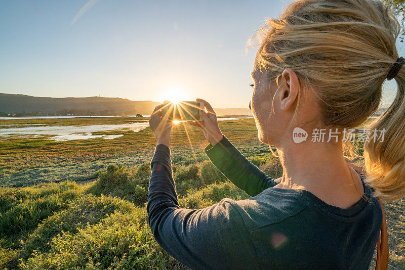 一名年轻女子用智能手机拍摄湿地上的日落