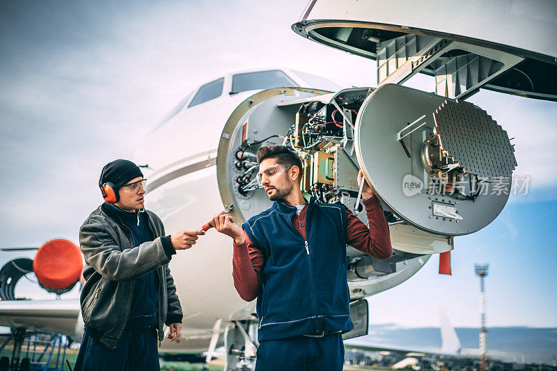 两名飞机机械师在一架停在机场的私人飞机前锥体下的雷达阵列上工作