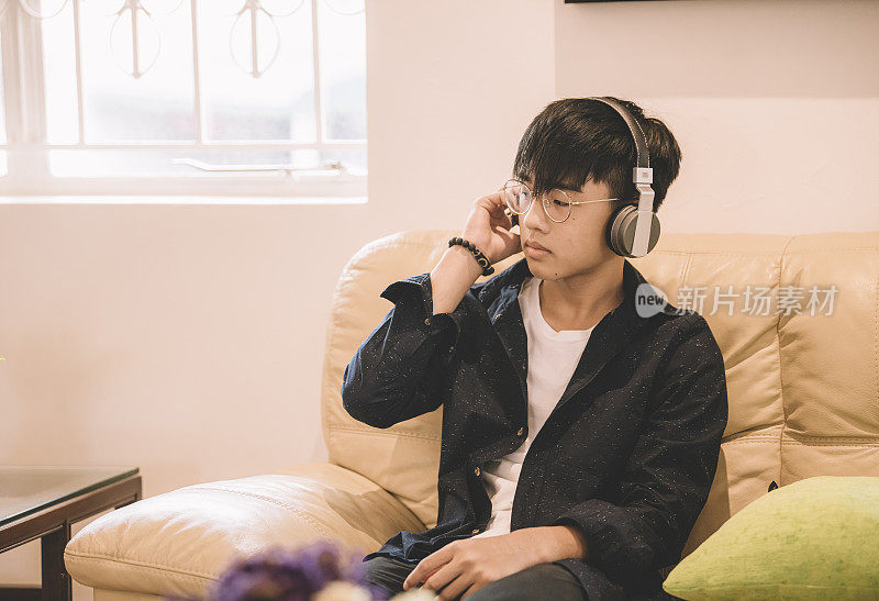 一个亚洲华裔少年周末坐在客厅的沙发上用耳机听音乐