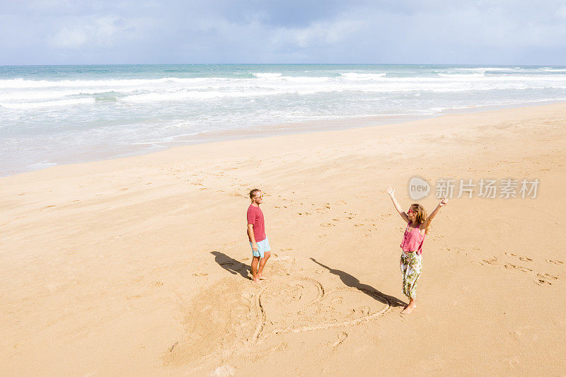 无人机上的年轻夫妇站在海滩上靠近画的心在沙滩上