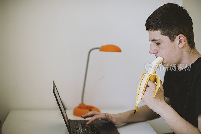 十几岁的男孩一边吃香蕉一边在电脑上做作业