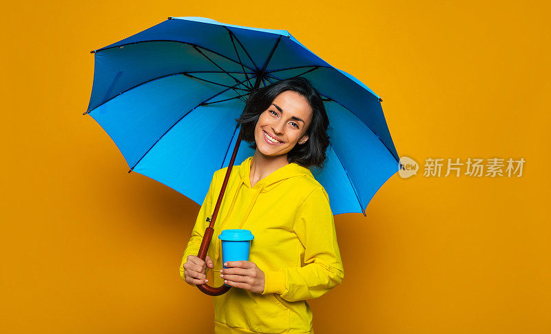 享受雨天。这是一张甜美女孩的半长照片，她撑着一把伞，面带微笑，穿着黄色的连帽衫，左手拿着蓝色的保温杯。