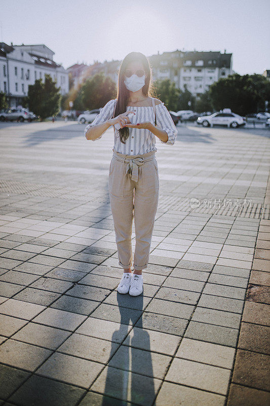 一名年轻女子戴着外科口罩，手上涂着洗手液，在封锁期间站在空荡荡的城镇广场上