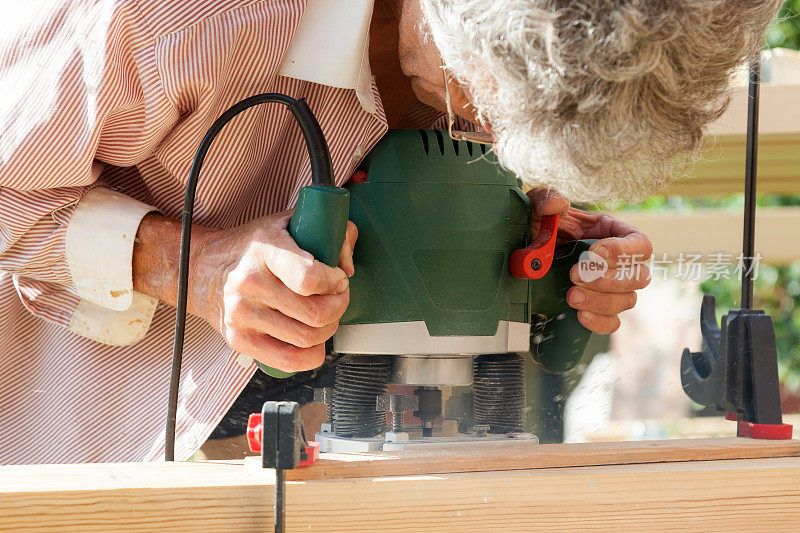 这位头发灰白的木匠手里拿着一个工具——一台绿色的铣床，在夏季小屋施工期间在一块新木板上切割。木工及木材加工