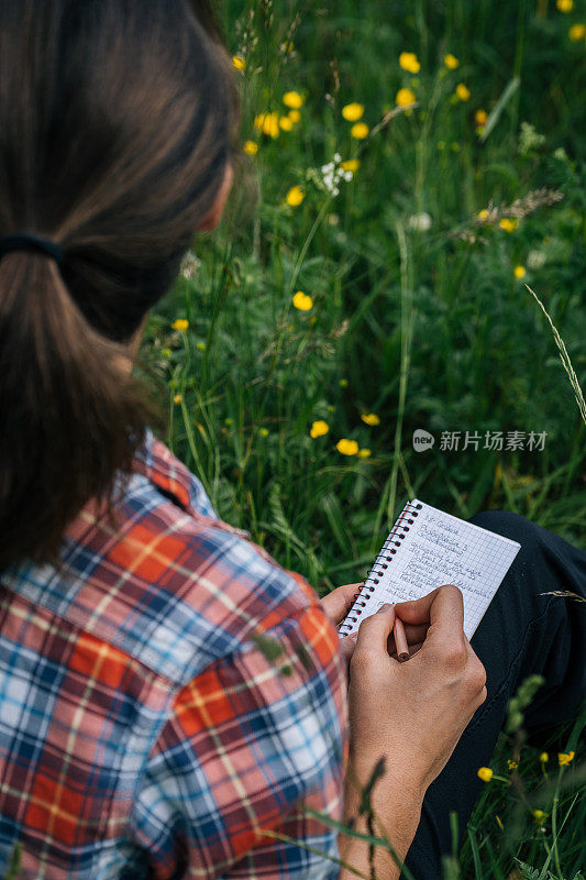 一个年轻的女人坐在山上的草地上，手里拿着笔记本和铅笔