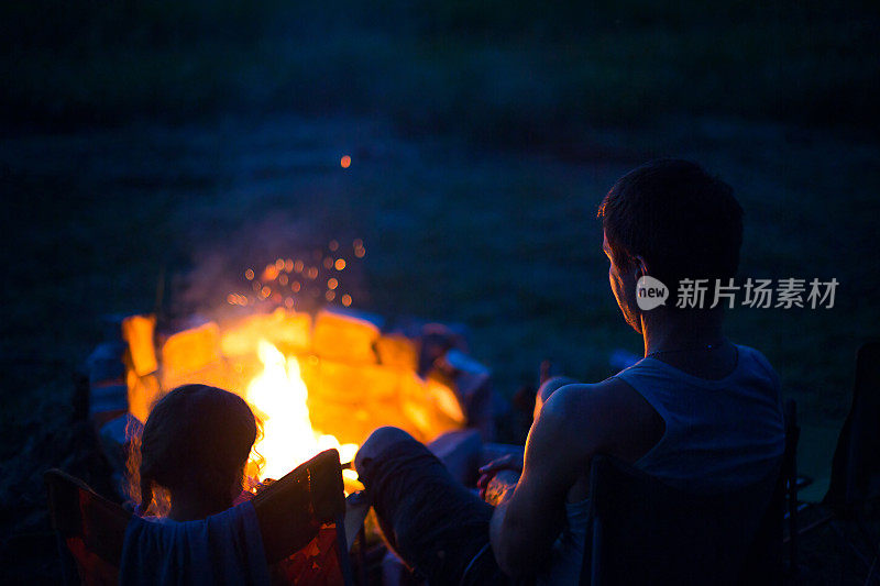 在夏天的大自然中，爸爸和女儿晚上坐在露天的火炉旁。家庭野营，围坐在篝火旁。父亲节,烧烤。野营灯和帐篷