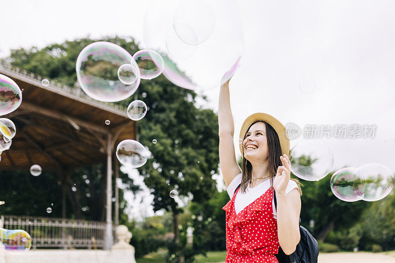 快乐的年轻西班牙妇女玩肥皂泡在城市公园的乐趣-微笑愉快的潮女游客享受她的暑假-旅游生活方式的概念