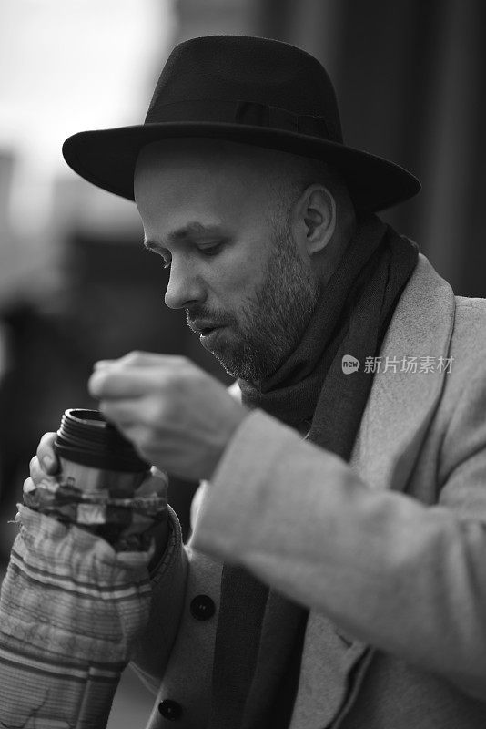 在莫斯科市中心，一名俄罗斯男子饿了，从热水瓶里吃东西。戴帽穿大衣的时髦男人。模糊的背景。黑白肖像。