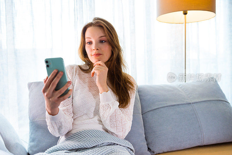 快乐的女人坐在沙发上用智能手机视频通话，托着下巴，听着。