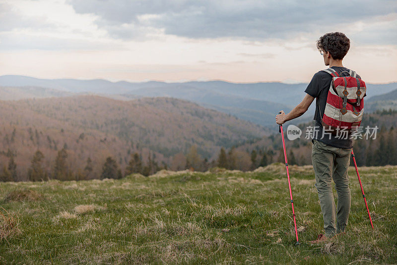 背着双肩包，拄着拐杖站在山顶的男旅行者的后面。