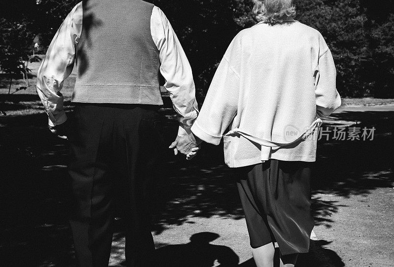 一对老夫妇在公园散步。祖母和祖父在金婚纪念日庆典上。50年的爱情故事。奶奶捧着花束，爷爷穿着背心牵着手。黑色和白色。