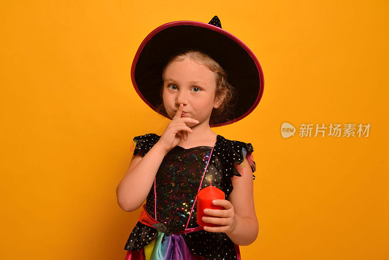 万圣节快乐。可爱的女孩在女巫服装显示沉默的手势或在黄色的背景上把手指放在嘴唇上。